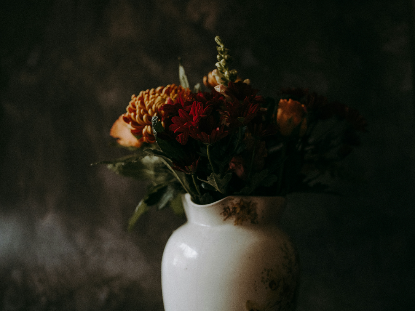 A bouquet of autumn flowers in a white ceramic vase, softly lit against a dark background